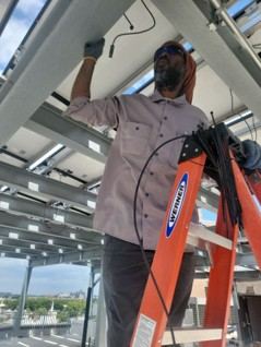 Technician on a ladder working on rooftop solar panel wiring with a city skyline in the background